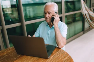 Idoso ao telefone resolvendo assuntos do INSS, com um laptop à frente, em casa