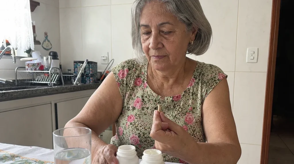 Idosa na cozinha segurando uma cápsula de suplemento, com copo de água e frascos na mesa