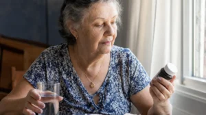 Idosa na luz da janela segurando um frasco de vitamina D e um copo de água